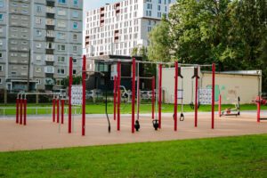 Āra trenažieru zona ar dažādiem vingrošanas rīkiem – pievilkšanās stieņiem, svarbumbām un pretestības gumijām; priekšplānā zaļa zāle, fonā daudzdzīvokļu ēkas un koki