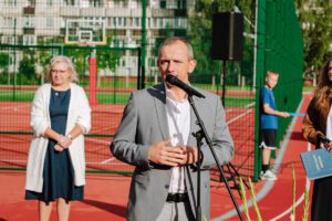 Vīrietis pelēkā uzvalkā runā mikrofonā āra pasākumā, fonā redzama sieviete baltā jaciņā un vēl viens cilvēks sporta tērpā pie žoga; pasākums notiek sporta laukumā vai līdzīgā vidē