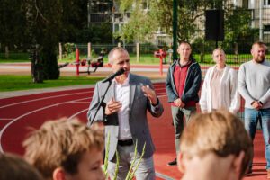 Cilvēku grupa stāv uz sarkanā skrejceļa; priekšplānā cilvēks uzvalkā runā mikrofonā un žestikulē, fonā redzami citi cilvēki, bērni un āra vingrošanas aprīkojums zaļā vidē.