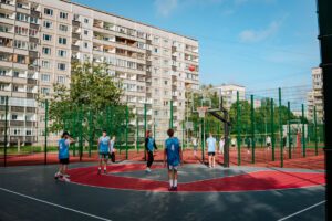 Jaunieši spēlē basketbolu uz āra laukuma, kas iežogots ar zaļu žogu; daļa tērpta zilās, daļa baltās sporta formās; fonā daudzdzīvokļu ēkas, skaidras debesis ar dažiem mākoņiem