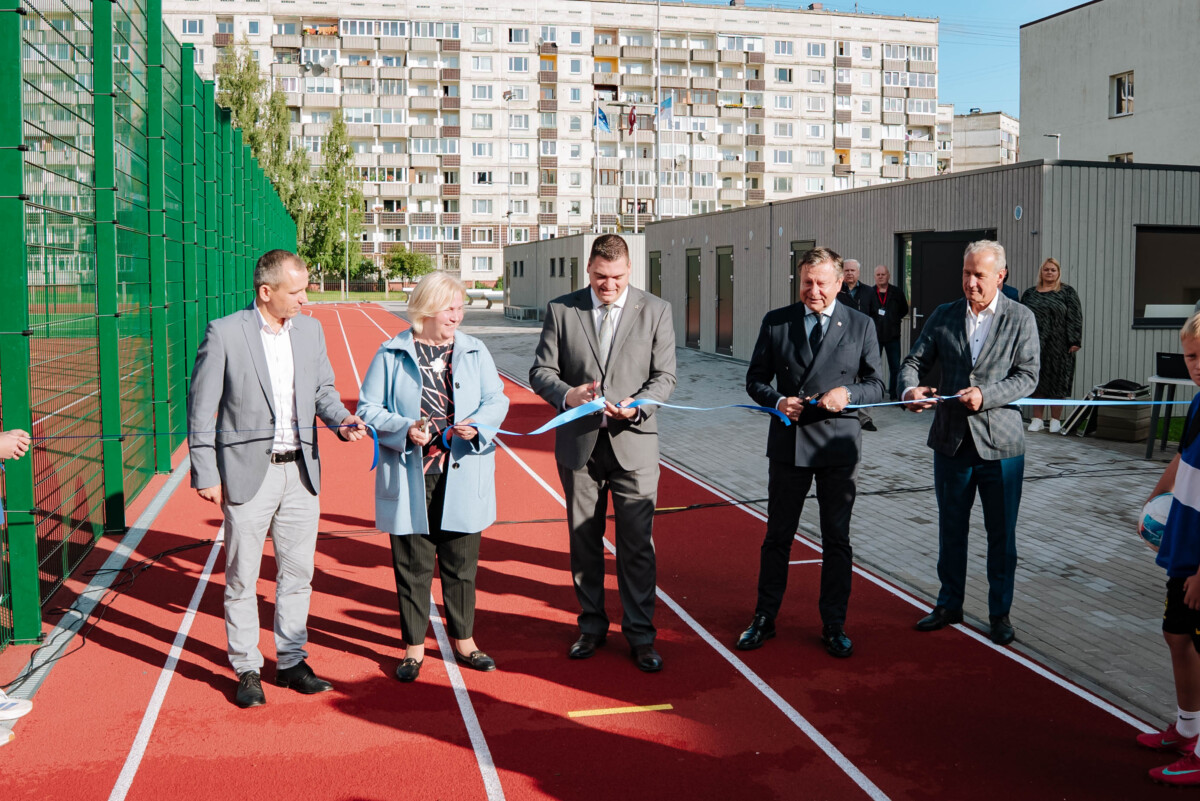 Pieci cilvēki stāv uz sarkanā skrejceļa un pārgriež zilu lenti, iespējams, atklāšanas ceremonijā; kreisajā pusē zaļš žogs, fonā ēkas