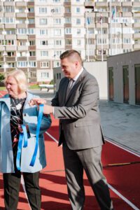 Divi cilvēki stāv uz sarkanā skrejceļa pie ēkas ar balkoniem, turot zilu lenti, iespējams, piedaloties atklāšanas ceremonijā; fonā redzamas karogu mastas un nelielas ēkas