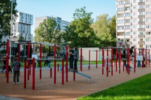 Āra rotaļu laukums ar sarkanām un sudrabotām kāpšanas konstrukcijām, horizontālajiem stieņiem un slidkalniņu; bērni rotaļājas, apkārt zaļa zāle, koki un daudzdzīvokļu ēkas