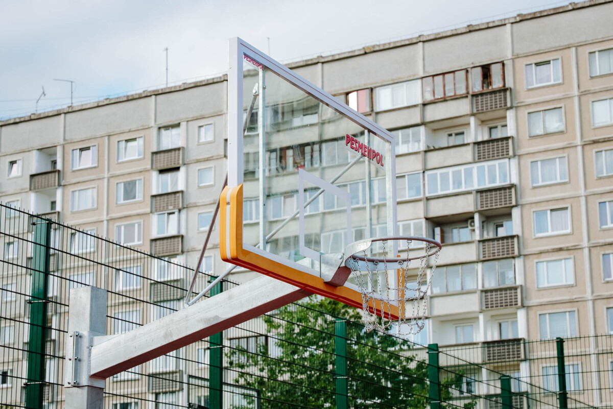 Āra basketbola grozs ar stikla vairogu uz metāla staba, laukums iežogots ar zaļu metāla žogu, fonā daudzdzīvokļu nams ar balkoniem, daļēji mākoņainas debesis