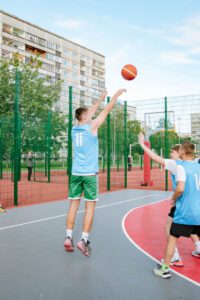 Jaunieši spēlē basketbolu uz āra laukuma ar sarkanu un pelēku segumu; spēlētājs ar 11. numuru met bumbu, bet spēlētājs ar 1. numuru mēģina bloķēt metienu; fonā redzami daudzdzīvokļu nami un koki