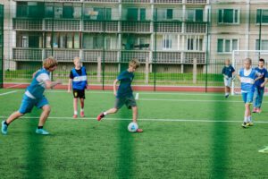 Bērni spēlē futbolu uz āra laukuma ar mākslīgo zālienu, laukums ir iežogots, fonā redzami daudzdzīvokļu nami ar balkoniem