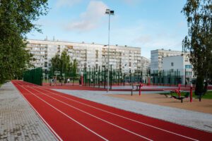 Āra sporta komplekss ar sarkanu skrejceļu, basketbola laukumiem un vingrošanas aprīkojumu, fonā redzami daudzdzīvokļu nami un koki, cilvēki aktīvi sporto, debesīs daļēji mākoņains