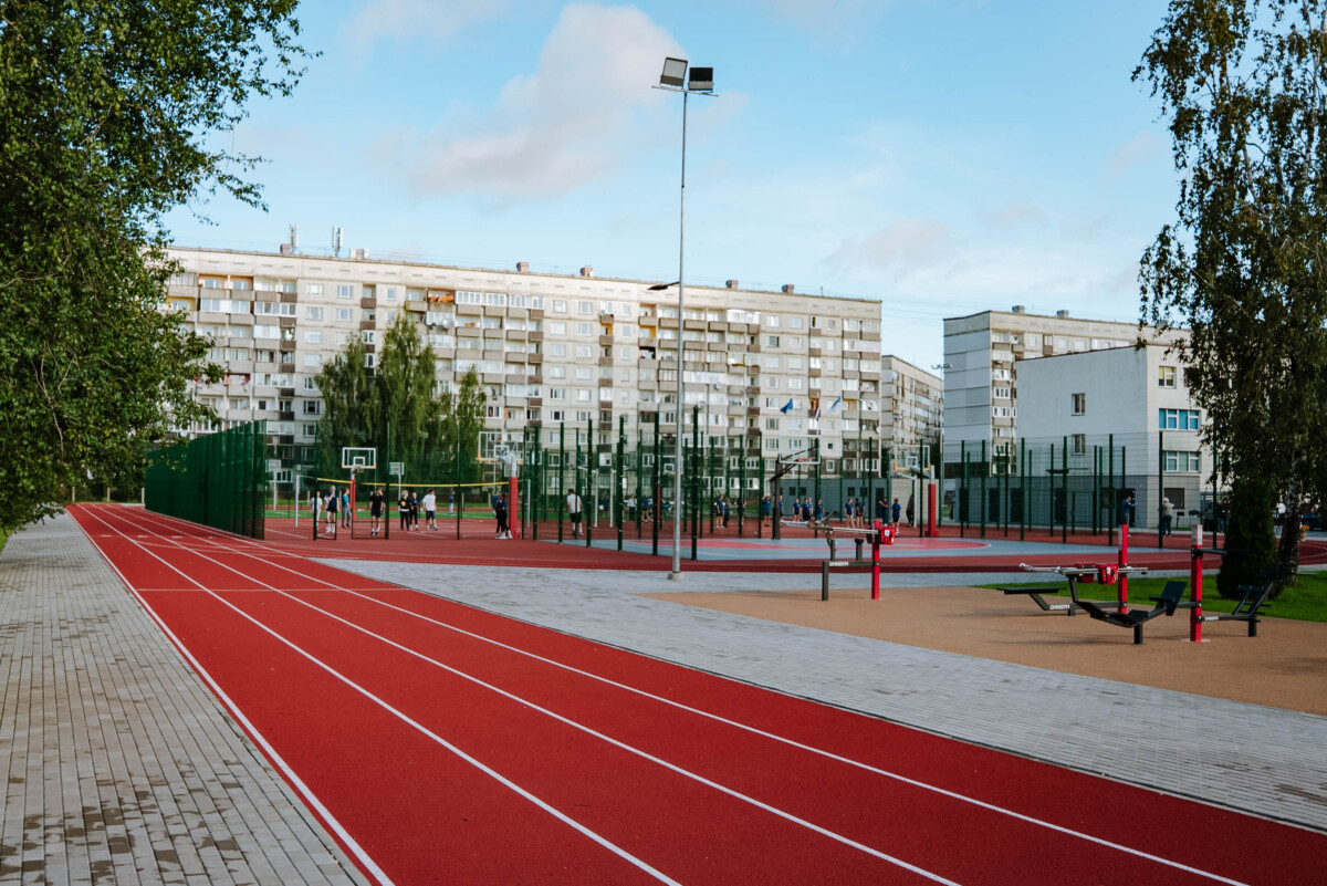 Āra sporta komplekss ar sarkanu skrejceļu, basketbola laukumiem un vingrošanas aprīkojumu, fonā redzami daudzdzīvokļu nami un koki, cilvēki aktīvi sporto, debesīs daļēji mākoņains