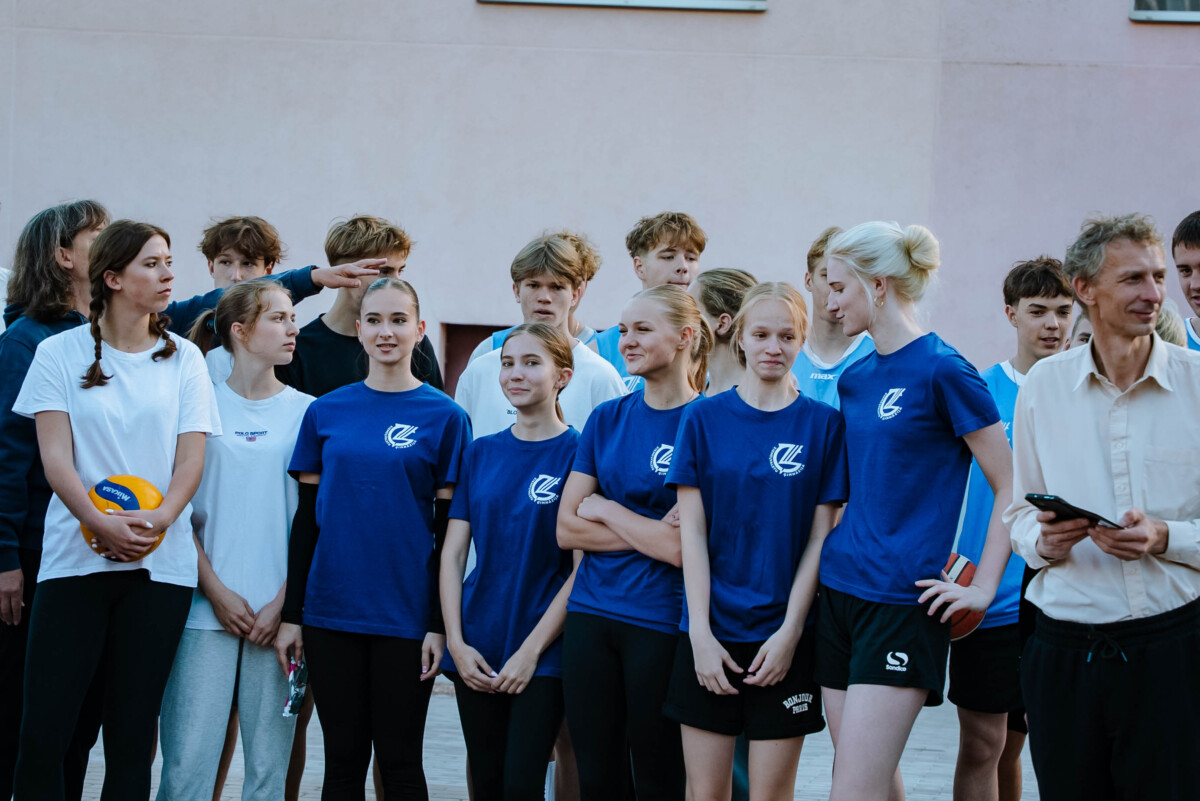 Cilvēku grupa stāv kopā, daļa tērpta zilos kreklos ar baltu logotipu, viens cilvēks tur volejbola bumbu