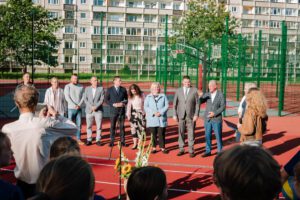 Cilvēku grupa stāv rindā uz sarkana āra sporta laukuma, iespējams, pasākuma vai ceremonijas laikā; daļa tērpta formālā un pusformālā apģērbā; fonā zaļš žogs, basketbola grozs un daudzdzīvokļu ēka ar balkoniem