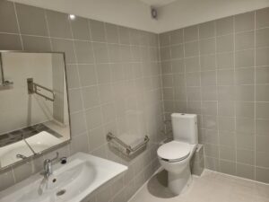 Tualēte ar gaiši pelēkām flīzēm uz sienām, balta izlietne ar krānu un spoguli kreisajā pusē, tualetes pods labajā pusē, pie sienas uzstādīti metāla rokturi