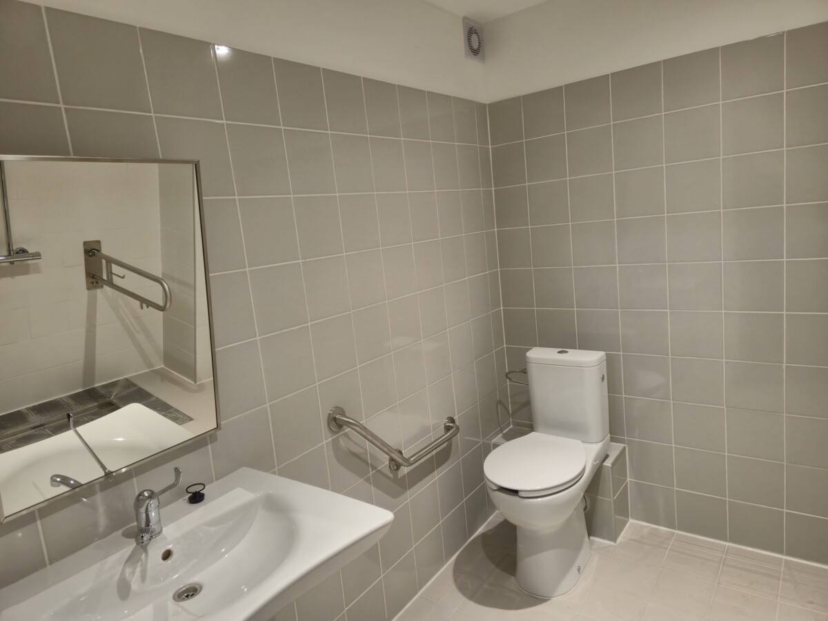 Tualēte ar gaiši pelēkām flīzēm uz sienām, balta izlietne ar krānu un spoguli kreisajā pusē, tualetes pods labajā pusē, pie sienas uzstādīti metāla rokturi