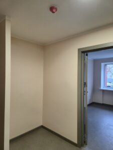 Neliela telpa ar gaišām sienām un pelēku grīdu, griestos uzstādīts sarkans dūmu detektors; atvērta durvju aile ved uz citu istabu ar logu