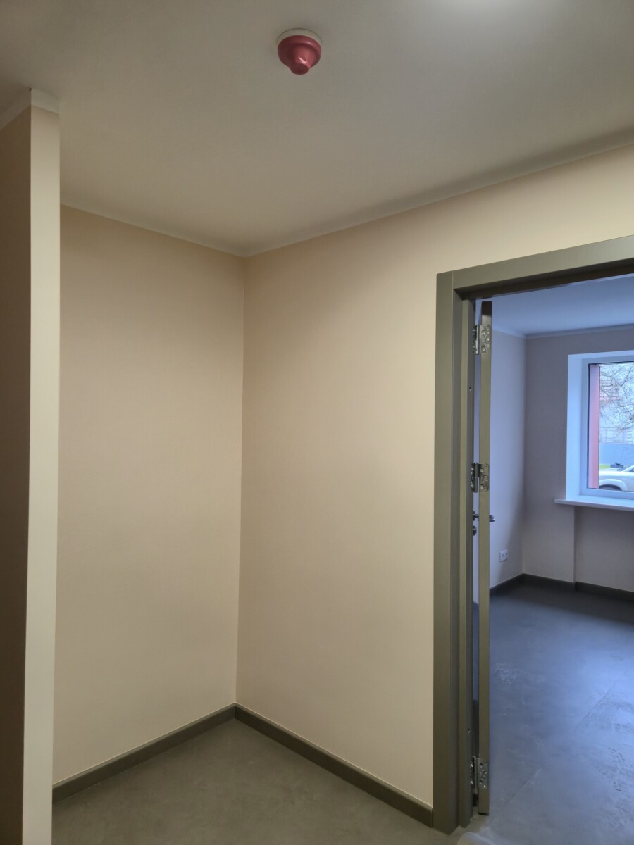 Neliela telpa ar gaišām sienām un pelēku grīdu, griestos uzstādīts sarkans dūmu detektors; atvērta durvju aile ved uz citu istabu ar logu