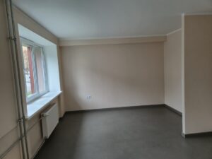 Tukša istaba ar gaišām sienām, lielu logu kreisajā pusē un radiatoru zem loga; grīda tumšā krāsā