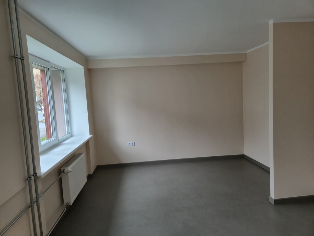 Tukša istaba ar gaišām sienām, lielu logu kreisajā pusē un radiatoru zem loga; grīda tumšā krāsā