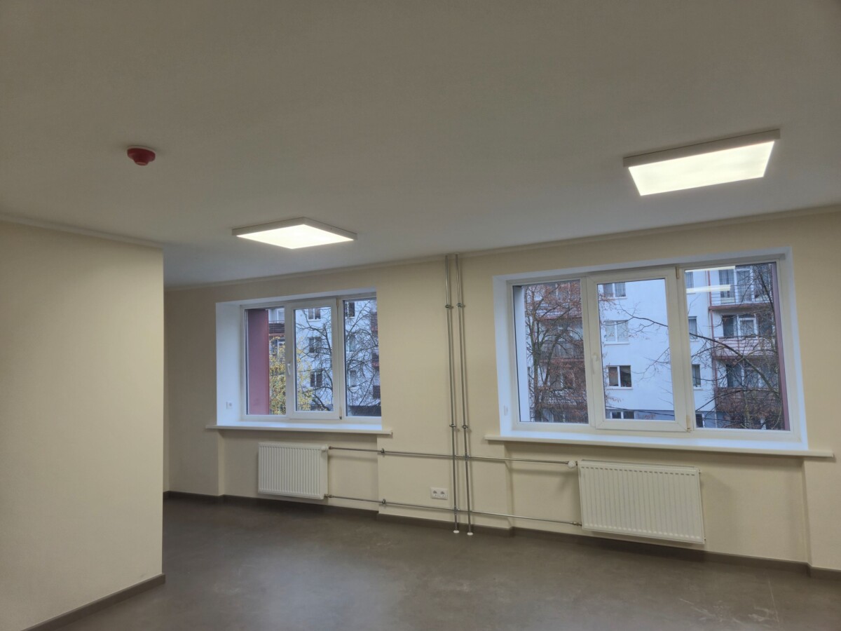 Plaša telpa ar gaišām sienām un tumšu grīdu, divi logi ar baltiem rāmjiem un radiatoriem zem tiem, griestos divi kvadrātveida gaismekļi un sarkans dūmu detektors