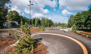 Asfalta pumptrack trase ar plūstošiem pagriezieniem un pacēlumiem, ko ieskauj dažādi apstādījumi — mazi skujkoki, krūmi un mulčas zonas. Fonā redzami koki, žogs un balta ēka, bet virs laukuma — zilas debesis ar baltiem mākoņiem un augsti apgaismes stabi.