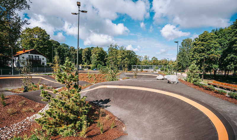 Asfalta pumptrack trase ar plūstošiem pagriezieniem un pacēlumiem, ko ieskauj dažādi apstādījumi — mazi skujkoki, krūmi un mulčas zonas. Fonā redzami koki, žogs un balta ēka, bet virs laukuma — zilas debesis ar baltiem mākoņiem un augsti apgaismes stabi.