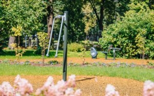 Āra vingrošanas laukums parkā ar vienkāršu metāla trenažieri priekšplānā, kas paredzēts roku un plecu kustību vingrinājumiem. Apkārt redzams zaļš zāliens, dekoratīvi krūmi, puķu dobes un koki, bet fonā – vēl citi āra sporta elementi. Skaidra, saulaina diena ar zilām debesīm un viegliem mākoņiem.