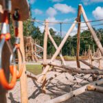 Koka rotaļu laukums ar dažādām līdzsvara un kāpelēšanas konstrukcijām, tostarp šķēršļu elementiem no koka stabiem, virvēm un tīkla kāpelēšanas zonām. Priekšplānā redzami koka elementi un metāla karabīņu stiprinājumi, bet fonā — lielāka konstrukcija ar metāla spirāles slidkalniņu. Apkārt redzami koki un plaša āra vide zem zilām debesīm ar mākoņiem.