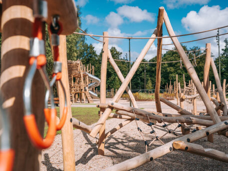 Koka rotaļu laukums ar dažādām līdzsvara un kāpelēšanas konstrukcijām, tostarp šķēršļu elementiem no koka stabiem, virvēm un tīkla kāpelēšanas zonām. Priekšplānā redzami koka elementi un metāla karabīņu stiprinājumi, bet fonā — lielāka konstrukcija ar metāla spirāles slidkalniņu. Apkārt redzami koki un plaša āra vide zem zilām debesīm ar mākoņiem.