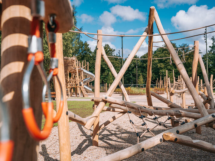 Koka rotaļu laukums ar dažādām līdzsvara un kāpelēšanas konstrukcijām, tostarp šķēršļu elementiem no koka stabiem, virvēm un tīkla kāpelēšanas zonām. Priekšplānā redzami koka elementi un metāla karabīņu stiprinājumi, bet fonā — lielāka konstrukcija ar metāla spirāles slidkalniņu. Apkārt redzami koki un plaša āra vide zem zilām debesīm ar mākoņiem.