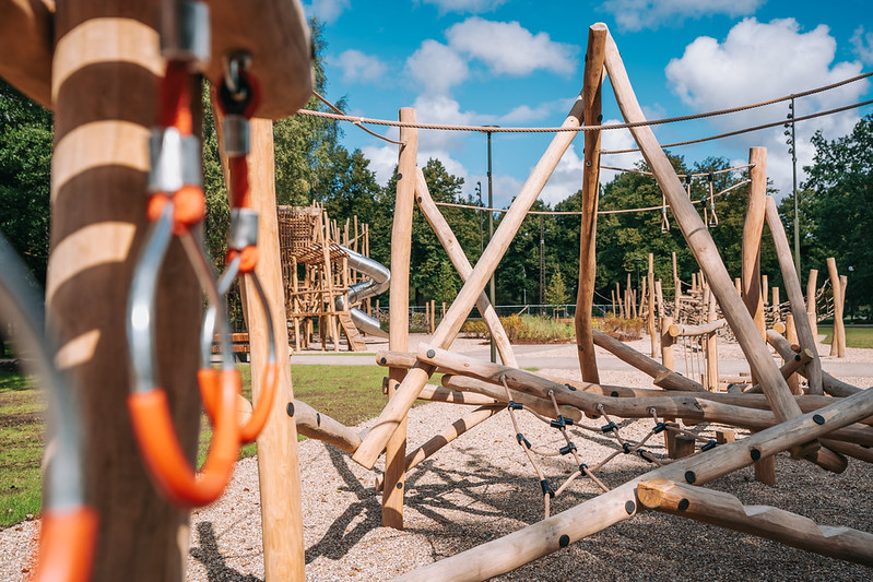 Koka rotaļu laukums ar dažādām līdzsvara un kāpelēšanas konstrukcijām, tostarp šķēršļu elementiem no koka stabiem, virvēm un tīkla kāpelēšanas zonām. Priekšplānā redzami koka elementi un metāla karabīņu stiprinājumi, bet fonā — lielāka konstrukcija ar metāla spirāles slidkalniņu. Apkārt redzami koki un plaša āra vide zem zilām debesīm ar mākoņiem.