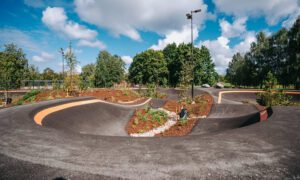 Asfalta pumptrack trase ar viļņotiem pagriezieniem un pacēlumiem, kuru ieskauj apzaļumoti stādījumi, mulčas zonas un jauni koki. Fonā redzams koku masīvs, zilas debesis ar mākoņiem un augsti apgaismes stabi gar trases malām.