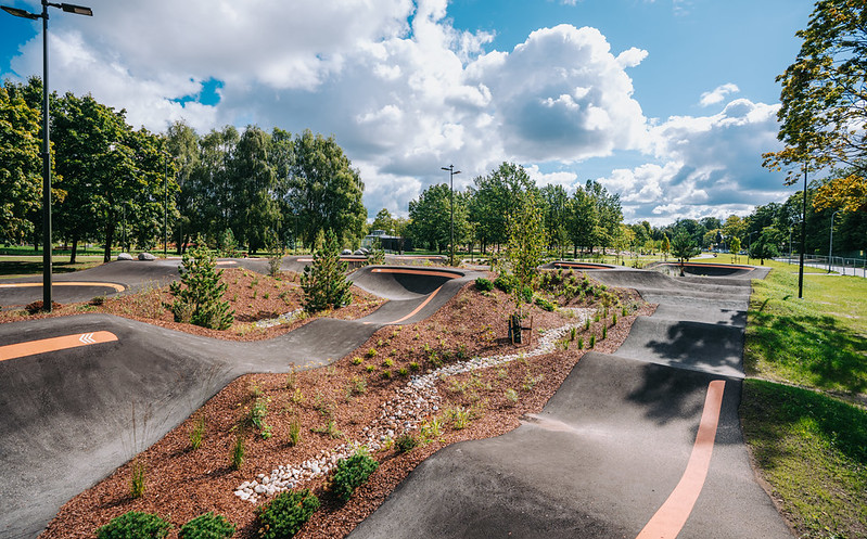 Plašs āra pumptrack laukums ar asfalta viļņiem un pagriezieniem, kuru ieskauj apstādījumi ar krūmiem, dekoratīviem stādiem un skujkokiem. Fonā redzami koki, zils debesu fons ar baltajiem mākoņiem un lampu stabi gar trases malām.