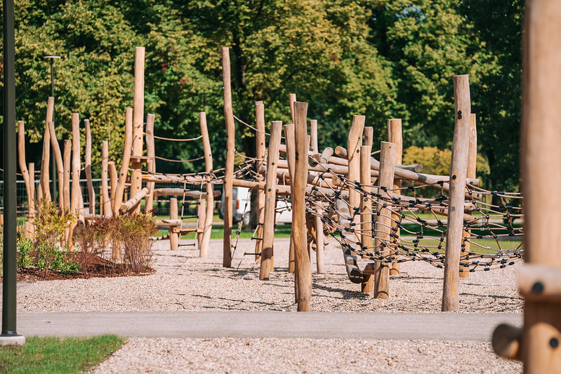 Koka rotaļu laukuma zona ar vertikāliem koka stabiem, virvju šķēršļiem un līdzsvara elementiem, izvietota uz grants seguma ar kokiem fonā
