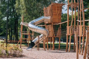 Bērnu rotaļu laukums ar lielām koka konstrukcijām, virvju tiltiņiem un metāla spirāles slidkalniņu. Apkārt ir koki un soliņi, bet zemē – smilšaina vai grants seguma zona.