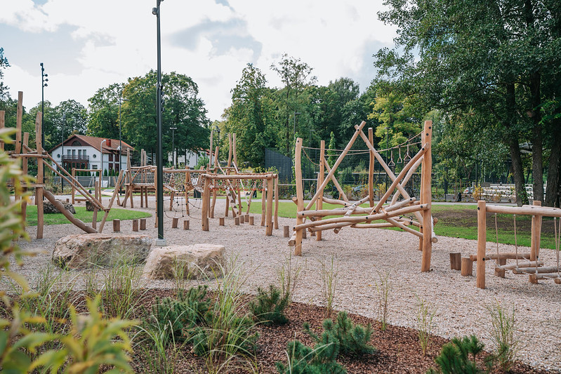 Koka rotaļu laukums ar dažādām kāpelēšanas un līdzsvara konstrukcijām, izvietots uz grants seguma. Redzami koka stabi, virvju elementi un horizontālas kāpšanas sijas, bet fonā – koki, ēkas un plaša parka vide