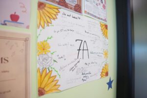 Uz gaiši zaļas sienas piestiprināts plakāts ar lielu uzrakstu “7A” vidū. Plakāta malās ir krāsotas saulespuķes un rozes, bet apkārt uzrakstam redzami dažādi ar roku rakstīti novēlējumi un īsi teikumi. Apkārt sienai redzami arī citi piekārti materiāli, tostarp sertifikāts un izglītojoši plakāti ar ilustrācijām. Kopējais iespaids — skolēnu veidots dekoratīvs apsveikuma vai klases plakāts.