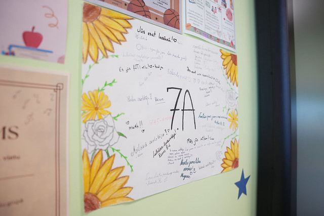 Uz gaiši zaļas sienas piestiprināts plakāts ar lielu uzrakstu “7A” vidū. Plakāta malās ir krāsotas saulespuķes un rozes, bet apkārt uzrakstam redzami dažādi ar roku rakstīti novēlējumi un īsi teikumi. Apkārt sienai redzami arī citi piekārti materiāli, tostarp sertifikāts un izglītojoši plakāti ar ilustrācijām. Kopējais iespaids — skolēnu veidots dekoratīvs apsveikuma vai klases plakāts.