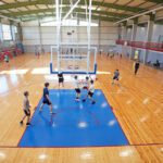 Plaša iekštelpu sporta zāle ar gaišu koka grīdu un zilu basketbola laukuma zonējumu. Centrā redzams basketbola grozs ar balta metāla konstrukcijām, kas karājas no griestiem. Apkārt laukumam vairākas cilvēku grupas trenējas vai spēlē basketbolu — daļa driblē, daļa met bumbu vai pārvietojas pa laukumu. Fonā gar labo pusi ir sarkanas margas un vairākas ieejas durvis, bet kreisajā pusē redzamas pelēkas un sarkanas tribīnes. Lieli logi sienas augšdaļā ielaiž daudz dienasgaismas, izgaismojot visu telpu.