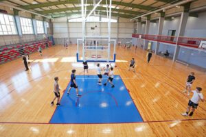 Plaša iekštelpu sporta zāle ar gaišu koka grīdu un zilu basketbola laukuma zonējumu. Centrā redzams basketbola grozs ar balta metāla konstrukcijām, kas karājas no griestiem. Apkārt laukumam vairākas cilvēku grupas trenējas vai spēlē basketbolu — daļa driblē, daļa met bumbu vai pārvietojas pa laukumu. Fonā gar labo pusi ir sarkanas margas un vairākas ieejas durvis, bet kreisajā pusē redzamas pelēkas un sarkanas tribīnes. Lieli logi sienas augšdaļā ielaiž daudz dienasgaismas, izgaismojot visu telpu.