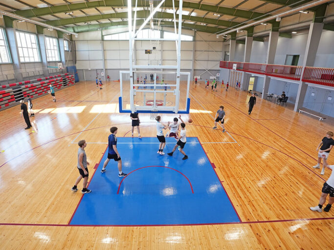 Plaša iekštelpu sporta zāle ar gaišu koka grīdu un zilu basketbola laukuma zonējumu. Centrā redzams basketbola grozs ar balta metāla konstrukcijām, kas karājas no griestiem. Apkārt laukumam vairākas cilvēku grupas trenējas vai spēlē basketbolu — daļa driblē, daļa met bumbu vai pārvietojas pa laukumu. Fonā gar labo pusi ir sarkanas margas un vairākas ieejas durvis, bet kreisajā pusē redzamas pelēkas un sarkanas tribīnes. Lieli logi sienas augšdaļā ielaiž daudz dienasgaismas, izgaismojot visu telpu.
