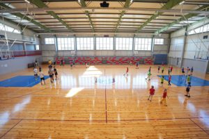 Plaša sporta zāle ar koka grīdu, kurā vienlaikus notiek vairākas aktivitātes. Dažādas vecuma grupas kustas pa laukumu, spēlējot basketbolu vai vingrinoties ar bumbām. Telpā ir augsti griesti ar metāla konstrukcijām, lieli logi gar vienu sienu ielaiž daudz dienasgaismas, bet otrā pusē redzamas sarkanpelēkas tribīnes. Laukuma centrā ir iezīmēts spēles logo, un ap zāli novietoti vairāki basketbola grozi.