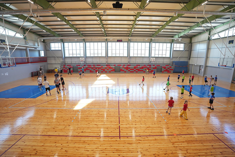 Plaša sporta zāle ar koka grīdu, kurā vienlaikus notiek vairākas aktivitātes. Dažādas vecuma grupas kustas pa laukumu, spēlējot basketbolu vai vingrinoties ar bumbām. Telpā ir augsti griesti ar metāla konstrukcijām, lieli logi gar vienu sienu ielaiž daudz dienasgaismas, bet otrā pusē redzamas sarkanpelēkas tribīnes. Laukuma centrā ir iezīmēts spēles logo, un ap zāli novietoti vairāki basketbola grozi.