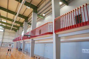 Plaša sporta zāles mala ar divu līmeņu konstrukciju. Apakšā redzama koka grīda un pelēkas sienas ar vairākām durvīm. Augšējā līmenī atrodas sarkanu metāla margu balkons, kas stiepjas gar visu sienu. No griestiem redzamas ventilācijas caurules un apgaismojums. Priekšplānā uz koka grīdas atrodas viena persona, kas tur sporta aprīkojumu. Kopējā vide ir gaiša un industriāli funkcionāla, raksturīga mūsdienīgai sporta ēkai.