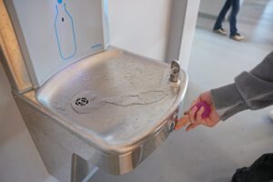 Nerūsējošā tērauda dzeramā ūdens strūklaka ar iebūvētu pudeles uzpildes paneli. Uz virsmas redzami ūdens šļaksti un notecējušas strūklas pēdas. Labajā pusē redzama cilvēka roka, kas nospiež pogu, lai ieslēgtu ūdens plūsmu.