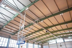 Sporta zāles griestu zona ar metāla sijām un paneļu konstrukcijām. No griestiem karājas basketbola groza pacelšanas mehānisms ar balta metāla rāmi un zilām detaļām. Attēla augšdaļā redzamas ventilācijas caurules un apgaismojuma lampas, bet zemāk — lieli logi, kas ielaiž spilgtu dienasgaismu. Kopējais skats parāda ēkas konstrukciju, tehnisko aprīkojumu un telpas plašumu.