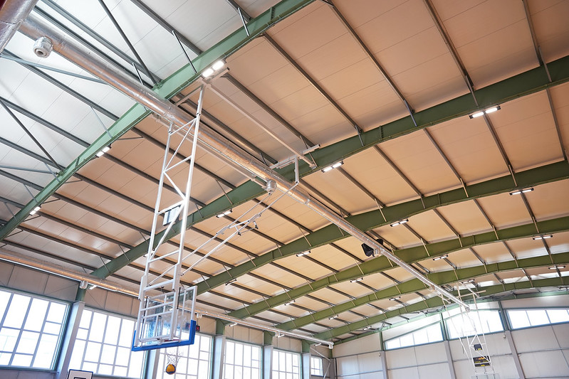 Sporta zāles griestu zona ar metāla sijām un paneļu konstrukcijām. No griestiem karājas basketbola groza pacelšanas mehānisms ar balta metāla rāmi un zilām detaļām. Attēla augšdaļā redzamas ventilācijas caurules un apgaismojuma lampas, bet zemāk — lieli logi, kas ielaiž spilgtu dienasgaismu. Kopējais skats parāda ēkas konstrukciju, tehnisko aprīkojumu un telpas plašumu.