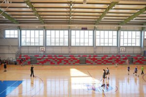 laša iekštelpu sporta zāle ar koka grīdu un augstiem logiem gar vienu sienu, kas ielaiž daudz dabiskās gaismas. Priekšplānā redzams basketbola laukuma centrālais apzīmējums, bet pa zāli pārvietojas vairāki cilvēki, daļa ar basketbola bumbām. Fonā atrodas sarkanpelēkas izbīdāmās tribīnes un vairāki basketbola grozi pie sienām. No griestiem karājas apgaismojuma un konstrukciju elementi, un telpas kopējais iespaids ir plašs un gaišs.