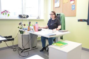 Gaišā, mājīgā kabinetā pie rakstāmgalda sēž persona, strādājot pie datora. Telpā ir liels logs, caur kuru ieplūst dienasgaisma, un uz palodzes novietots puķu pods. Uz galda atrodas ūdens pudele, kancelejas piederumi, dokumenti un dators. Blakus galdam ir neliels skapītis ar grāmatām, bet aiz personas redzams pie sienas piestiprināts ziņojumu dēlis. Kopējais iespaids — kārtīgs darba kabinets izglītības iestādē vai biroja vidē.