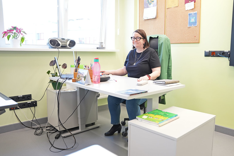 Gaišā, mājīgā kabinetā pie rakstāmgalda sēž persona, strādājot pie datora. Telpā ir liels logs, caur kuru ieplūst dienasgaisma, un uz palodzes novietots puķu pods. Uz galda atrodas ūdens pudele, kancelejas piederumi, dokumenti un dators. Blakus galdam ir neliels skapītis ar grāmatām, bet aiz personas redzams pie sienas piestiprināts ziņojumu dēlis. Kopējais iespaids — kārtīgs darba kabinets izglītības iestādē vai biroja vidē.