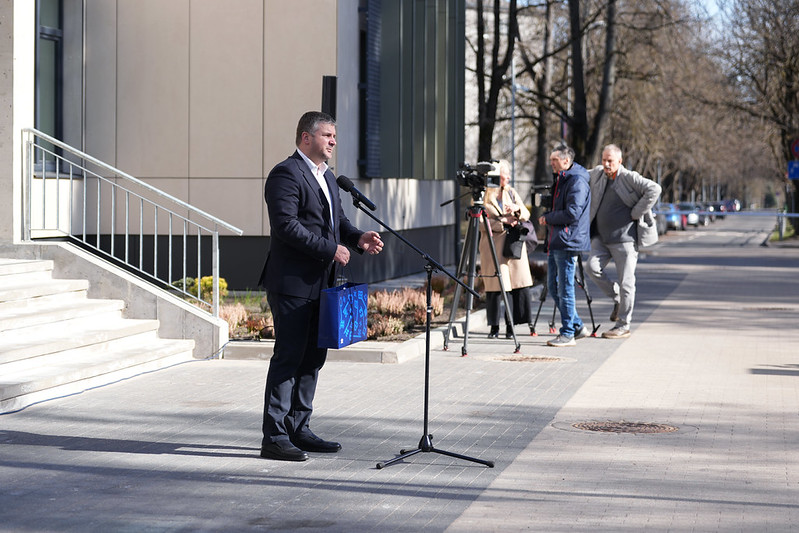 Persona uzstājas ar uzrunu pie ēkas, fonā mediju pārstāvji.