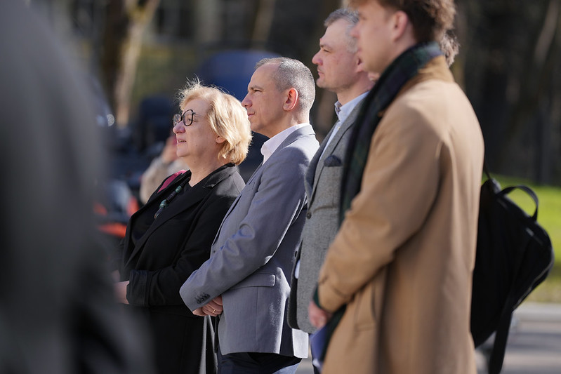 Pasākuma apmeklētāji klausās uzrunu āra teritorijā.