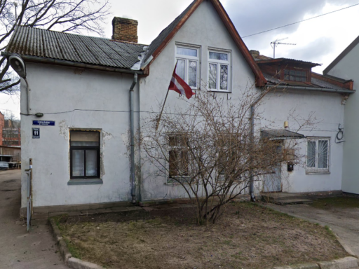 Divstāvu dzīvojamās ēkas foto Čiekurkalna 3. šķērslīnijā 11, Rīgā. Ēkai ir gaiši pelēks fasādes apmetums, slīps jumts un Latvijas karogs pie loga. Priekšā redzams neliels pagalms ar koku un bruģētu celiņu.